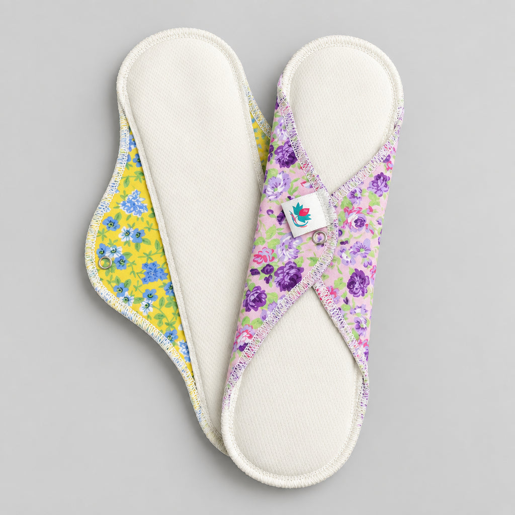 Deux serviettes hygiéniques de 11 1/2 pouces avec dessus en AWJ (tissu technique qui sèche rapidement). Une est jaune avec fleurs bleues et l'autre lilas avec fleurs roses et violettes