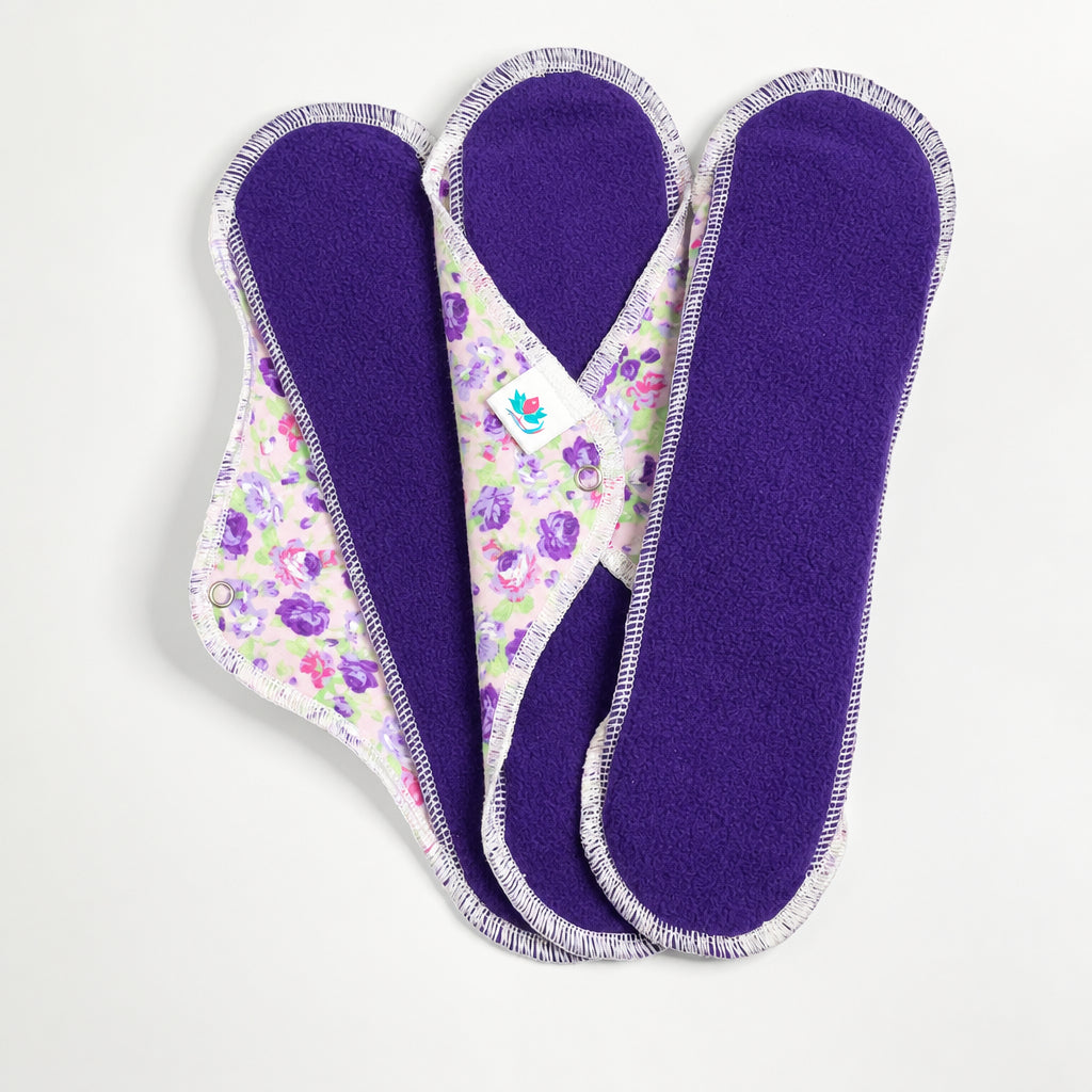 Serviettes hygiéniques extra longues en micropolar violet sur le dessus et flanelle de coton fleuri dans les tons de mauve et rose.